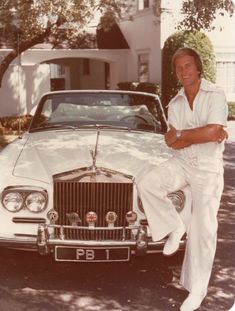 a man sitting on the hood of a white car in front of a house with his arms crossed