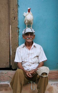 an old man with a chicken on his head sitting in front of a blue wall