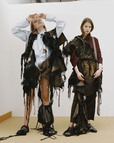 two women standing next to each other in front of a white wall with black fringes