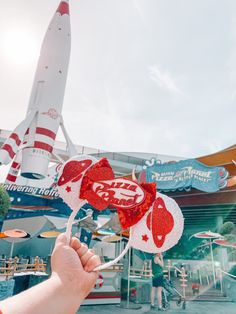 a hand holding up two red and white mouse ears in front of a rocket ship