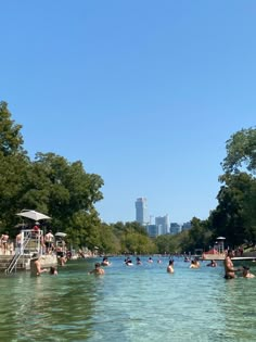 many people are swimming in the water near some trees and buildings on a sunny day