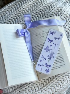 an open book with a blue ribbon on top of it next to a purple bow