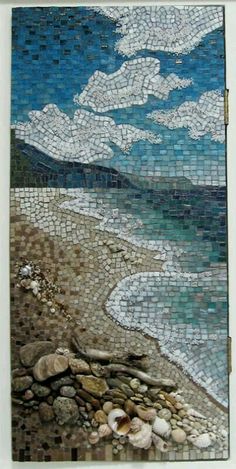 a mosaic tile wall hanging on the side of a building next to the ocean with rocks and pebbles