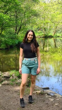 a woman standing in front of a river with trees and rocks on the bank behind her