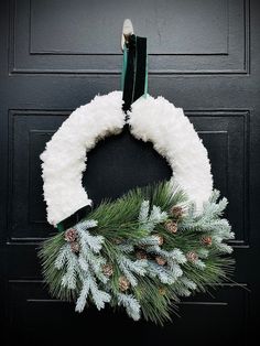 a white wreath with pine cones and evergreen needles hanging on a black front door frame