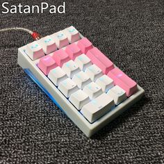 a pink and white computer keyboard sitting on top of a black carpeted floor with the caption satanpadd above it