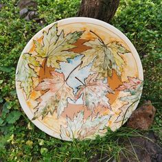 a plate with leaves painted on it sitting in the grass next to a tree trunk