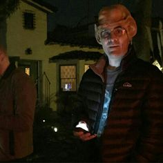 two men standing in front of a house at night, one holding a cell phone