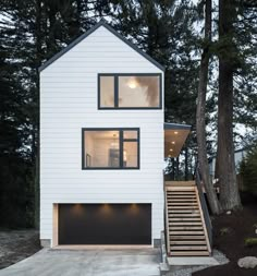 a white house with black windows and wooden steps leading up to the front door is surrounded by pine trees