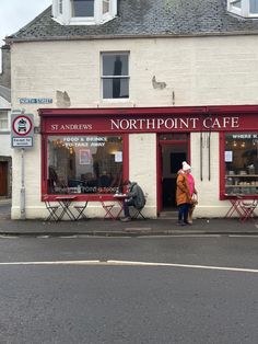 a woman standing in front of a store on the side of a road with tables and chairs outside