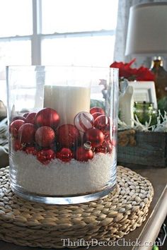 a candle sits in a glass container filled with christmas ornaments and white candlesticks