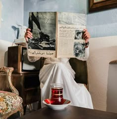 a person sitting at a table reading a book and holding up a newspaper with a cup of tea in front of them