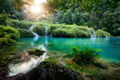 the sun shines brightly in front of a blue lake with waterfall and green vegetation