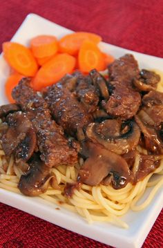 a white plate topped with meat and noodles next to carrots on a red table cloth