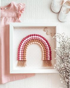 a pink and white framed artwork with a wooden rainbow in the middle next to baby's shoes