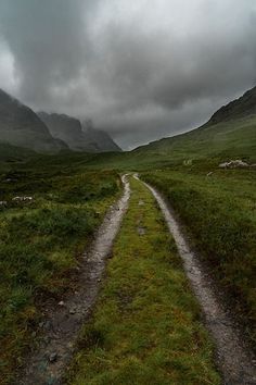 a dirt road in the middle of a grassy field with mountains in the background on a cloudy day