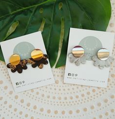 two pairs of earrings sitting on top of a table next to a green plant leaf