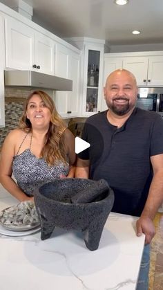 a man and woman standing in front of a bowl on a kitchen counter with white cabinets