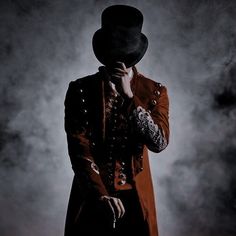 a man in a top hat and coat holding his hands to his face while standing against a dark background