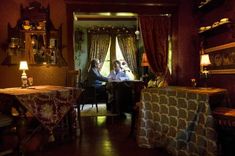 two people sitting at a table in a room with red walls and curtains on the windows