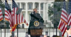 a man standing at a podium in front of american flags and talking on a cell phone