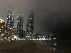 two people walking on the beach in front of some tall buildings with lights at night