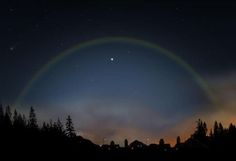 a rainbow in the night sky with trees and buildings behind it, all lit up