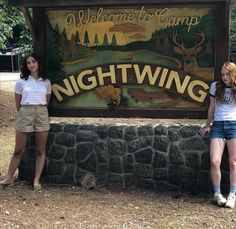 two girls standing in front of a sign for the nightwing campground, with trees and mountains in the background