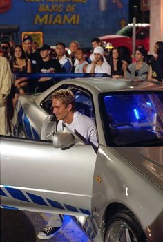 a man driving a silver sports car in front of a group of people on the street