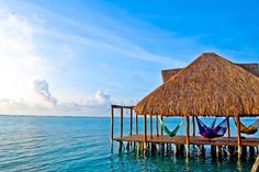 two hammocks sitting on a wooden dock in the water near a hut with thatched roof