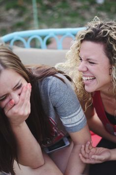 two women are laughing and sitting on the ground together, one is covering her face with her hand