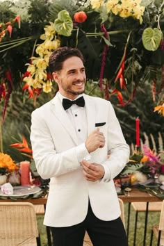 a man in a white tuxedo standing next to a table with flowers and candles