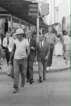 black and white photograph of men walking down the street in front of stores on a busy city street