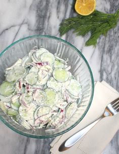 a glass bowl filled with cucumber salad next to a knife and fork on a marble surface