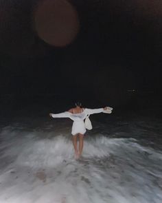 a woman standing in the ocean at night with her arms spread out and she is wearing a white suit