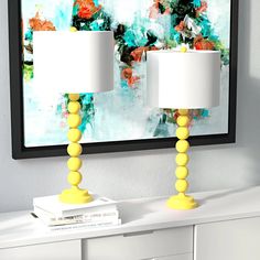 two yellow lamps sitting on top of a white dresser in front of a framed painting