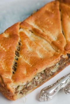 a piece of meat and cheese pie on a plate next to two silver spoons