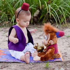 a baby sitting on a towel next to a stuffed animal and a toy monkey with a bell in its mouth