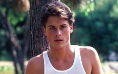 a young man standing next to a tree wearing a white tank top and holding a tennis racquet