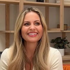 a woman with long blonde hair sitting in front of bookshelves and smiling at the camera