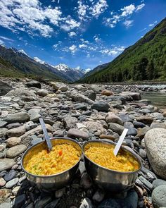 two metal bowls filled with food sitting on top of a rocky river bank next to mountains