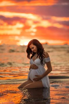 a pregnant woman sitting on the beach at sunset with her belly in the water while she is wearing a white dress