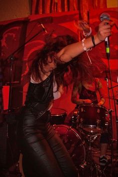 a woman in black leather pants holding a microphone and standing next to a drum set