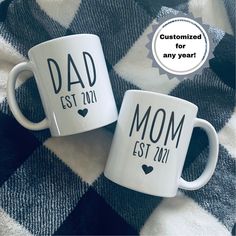 two coffee mugs with the words dad and mom on them sitting on a blanket