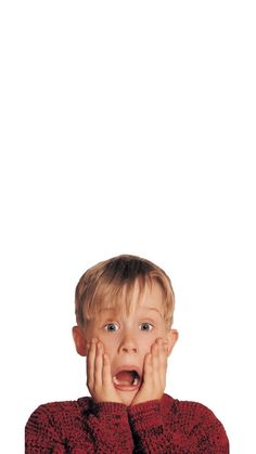 a young boy with his hands on his face looking at the camera while wearing a red sweater