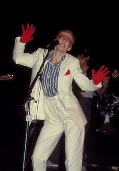 a man in a white suit and red gloves on stage with his hands out to the side