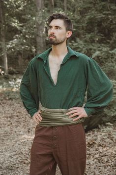 a man standing in the woods with his hands on his hips wearing brown pants and a green shirt