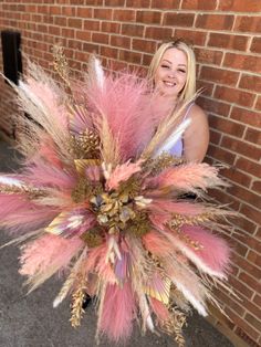 a woman standing next to a brick wall holding a large pink and gold feather bouquet