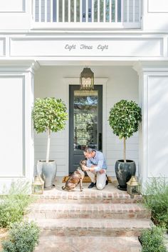 a man sitting on the steps with his dog in front of a white house that is decorated with potted trees