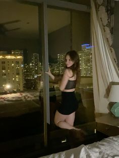 a woman standing in front of a window next to a bed and night time cityscape